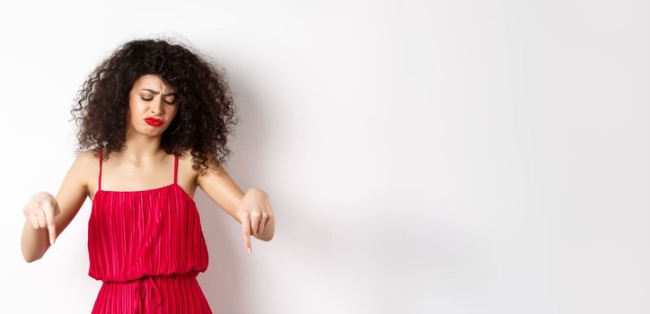 Sad and disappointed curly-haired woman in red dress, frowning and looking down jealous, pointing at promo, standing over white background.