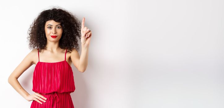 Smiling caucasian woman with curly hair, wearing red dress, showing rule number one gesture, raising finger and looking confident at camera, white background.