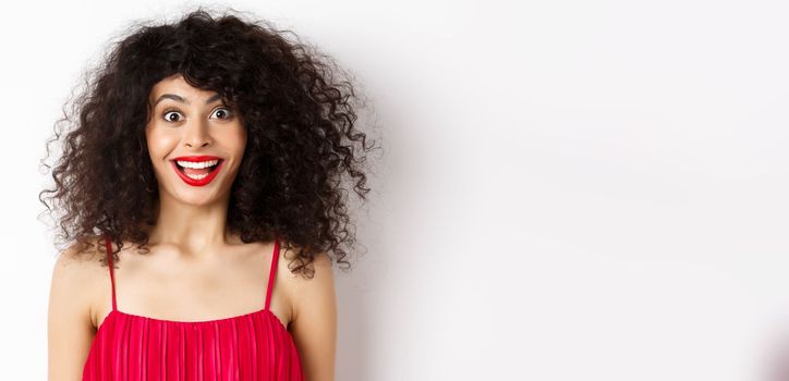 Close-up of happy lady with curly hair and red lips, raising eyebrows and looking surprised at camera, standing over white background.