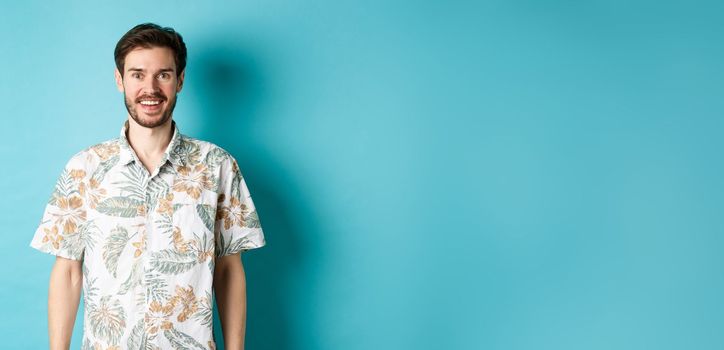 Cheerful caucasian guy going on vacation, wearing summer shirt and smiling, standing on blue background.