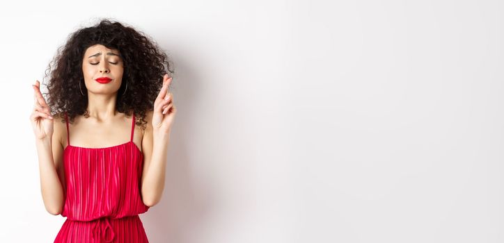 Hopeful woman in red dress making wish, praying with fingers crossed and closed eyes, pleading god, standing over white background.