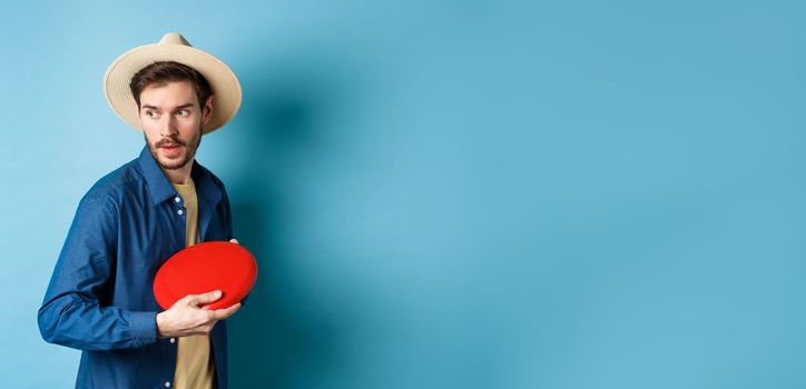 Handsome young guy in summer hat playing frisbee, throwing it aside, standing on blue background.