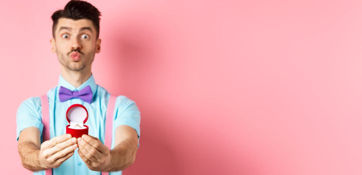 Valentines day. Funny young man pucker lips for kiss and showing engagement ring, making proposal, say marry me to lover, standing over pink background.