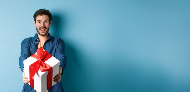 Valentines day. Excited handsome man giving gift box and wishing happy holiday. Romantic guy extending hands with present, standing againt blue background.