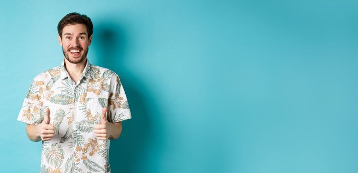 Summer holiday. Excited smiling tourist in hawaiian shirt show thumbs-up, praising hotel or vacation place, standing on blue background.
