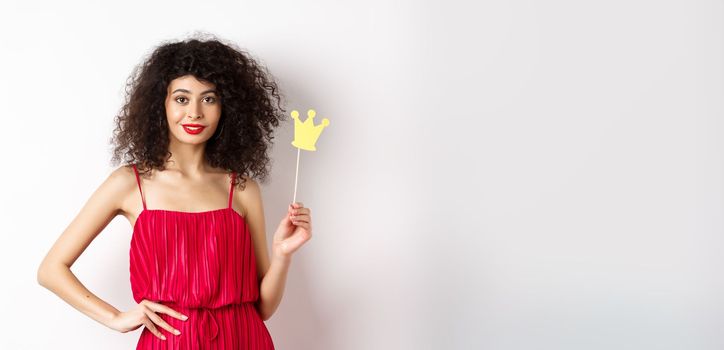 Stylish young woman in red dress, feeling confident and sassy, holding crown and smiling, standing over white background.