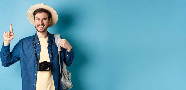 Handsome happy tourist in summer hat, holding backpack and camera, pointing finger up at logo, recommending travel agency or place on vacation, blue background.