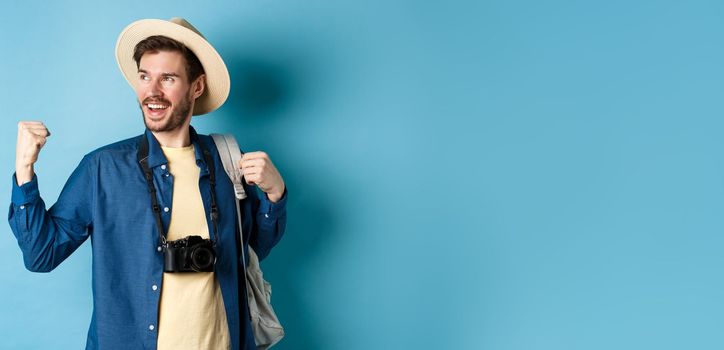 Cheerful tourist rejoicing on vacation, celebrating summer holidays, showing fist pump gesture and saying yes with satisfied face, looking aside, blue background.