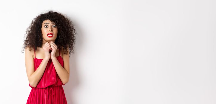 Scared caucasian woman trembling from fear, wearing red dress and staring anxious at camera, standing over white background.