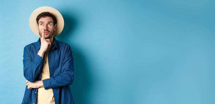 Thoughtful male tourist thinking of summer holiday vacation, looking aside with pensive face, touching lip and pondering next travel, standing on blue background.