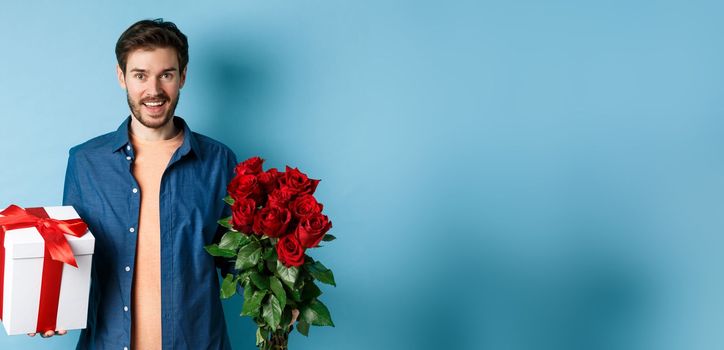 Love and relationship concept. Happy young man bring flowers and gift on romantic date. Boyfriend wish bouquet of roses and present standing on blue background.