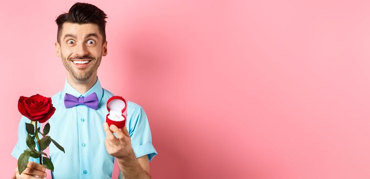 Valentines day. Smiling handsome man asking to marry him, showing engagement ring and red rose, standing romantic on pink background.