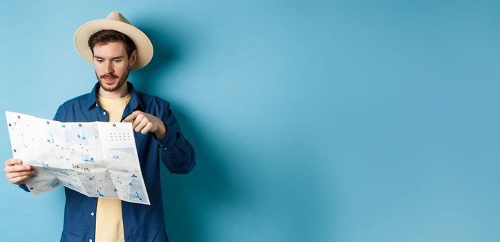 Handsome guy choosing place to go, pointing at sighseeing map on vacation, travelling on summer holidays, wearing straw hat and shirt, blue background.