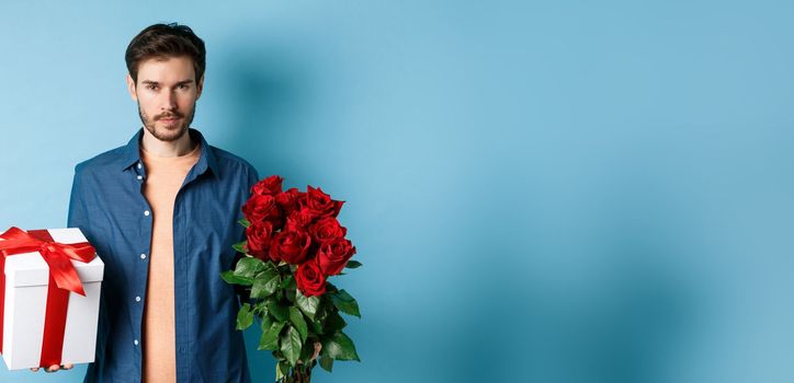 Love and Valentine day concept. Passionate man looking confident at camera, holding gift box and red roses for romantic date, standing over blue background.