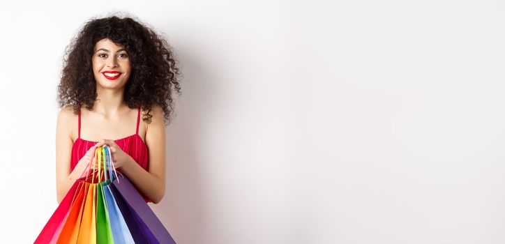 Image of stylish caucasian woman in red dress and makeup, holding shopping bags and smiling, standing over white background.