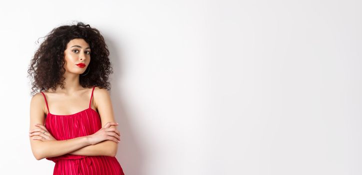 Sassy woman with curly hairstyle, wearing red dress and makeup, cross arms on chest, standing over white background.