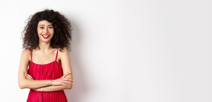 Elegant young woman in red dress with makeup, dressed-up for festive event, smiling happy at camera, standing over white background.