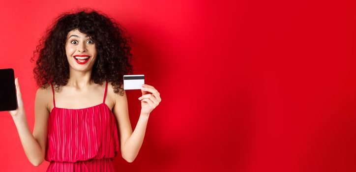 Excited lady in red dress showing plastic credit card and smartphone screen, smiling amazed, standing on studio background.