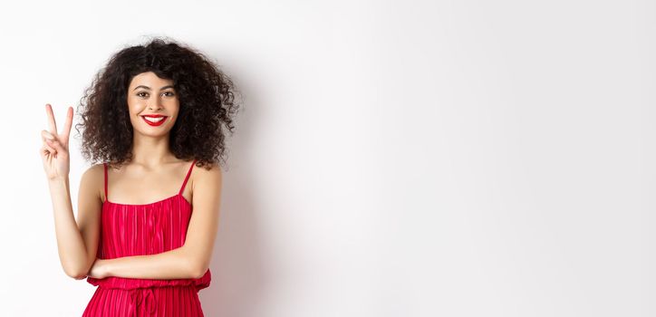 Attractive female model in red dress and makeup, showing number two and smiling, standing over white background.