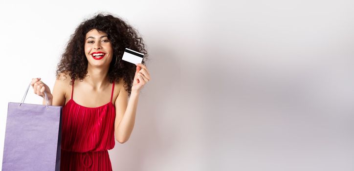 Beautiful woman with curly hair, red dress, showing shopping bag and plastic credit card, white background.