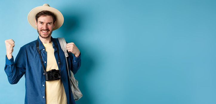 Happy tourist enjoying summer vacation, smiling and saying yes with fist pump, celebrating on holidays, feeling joy as finaly travel abroad, standing on blue background.