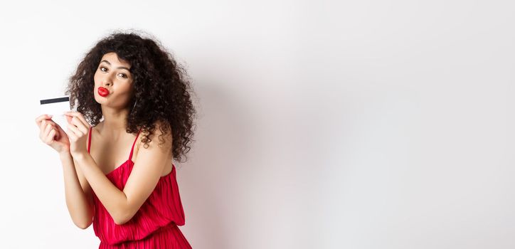 Shopping. Beautiful lady with curly hair, pucker lips, kissing plastic credit card, standing in red dress against white background.