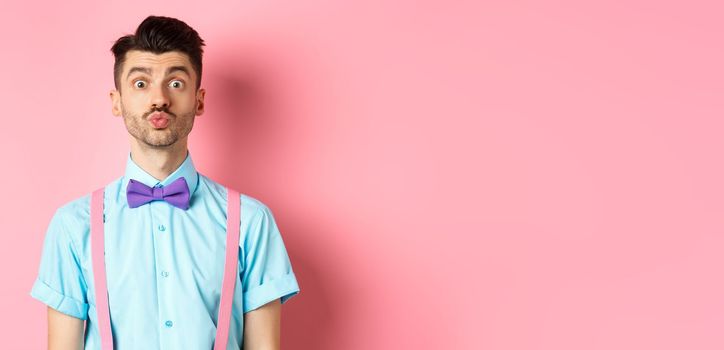 Funny young man waiting for kiss with puckered lips and silly face, standing on romantic pink background. Concept of love and Valentines day.