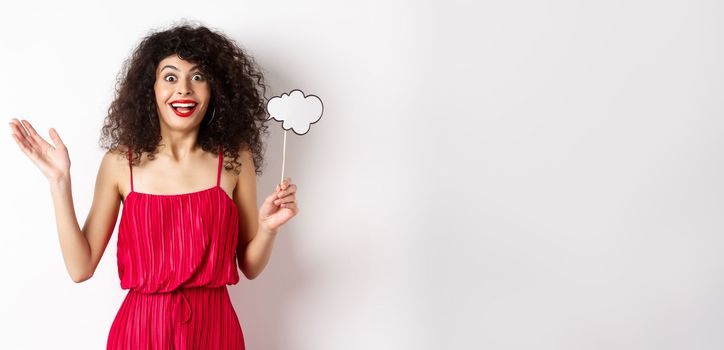 Happy young woman in red dress, holding comment cloud and looking surprised, standing over white background. Copy space