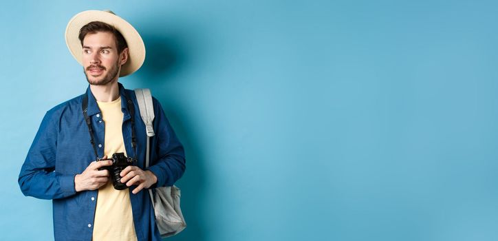 Handsome positive guy in summer hat, holding photo camera and looking aside, tourist taking pictures on vacation, standing with backpack on blue background.