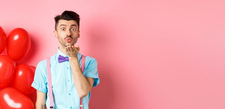 Valentines day concept. Attractive and funny man in bow-tie, blowing air kiss to lover, standing near pink background and red hearts balloons.