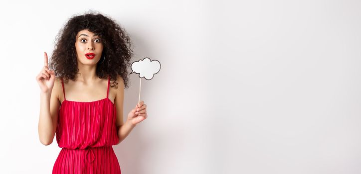 Cute caucasian lady in red dress, holding comment cloud and raising finger, pitching an idea and smiling, suggesting something, standing over white background.