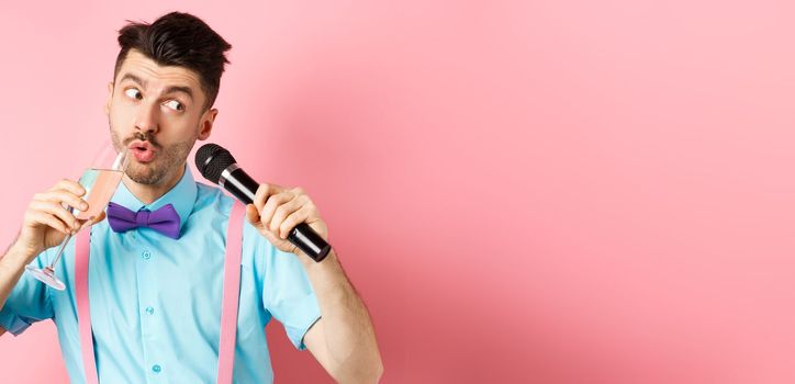 Party and festive events concept. Funny guy singing karaoke, performing song with microphone and drinking champagne from glass, standing on pink background.