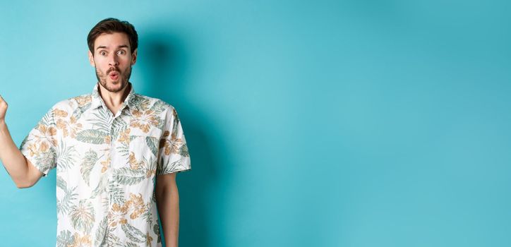 Surprised and amazed tourist in hawaiian shirt pointing finger left, showing promo and staring at camera, standing on blue background.