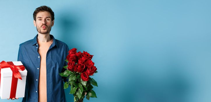 Love and Valentines day concept. Romantic boyfriend pucker lips for kiss, bring bouquet of red roses and gifts on date, standing over blue background.