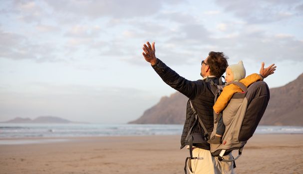 Father rising hands to the sky while enjoying pure nature carrying his infant baby boy son in backpack on windy sandy beach of Famara, Lanzarote island, Spain at sunset. Family travel concept