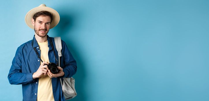 Handsome guy going on summer vacation, backpacking on holiday. Tourist with straw hat and camera smiling happy, standing on blue background.