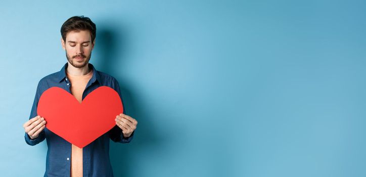 Lonely guy looking sad at valentines red heart with sad face, standing over blue background.