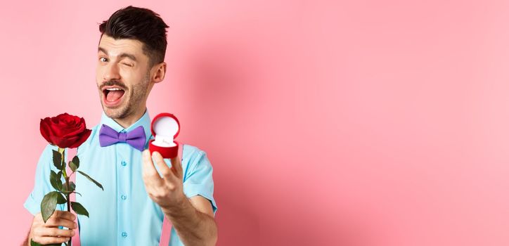 Valentines day. Funny guy making proposal, winking and saying marry me, showing engagement ring with red rose, standing over pink background.