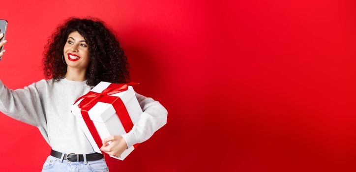 Happy young woman taking selfie with her valentines day gift, holding present and photographing on smartphone, posing on red background.