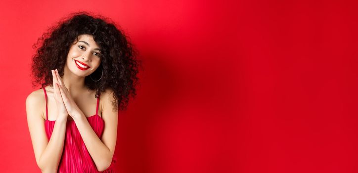 Tender woman with curly hair, wearing red dress, looking at something lovely, thanking you, standing over studio background. Copy space