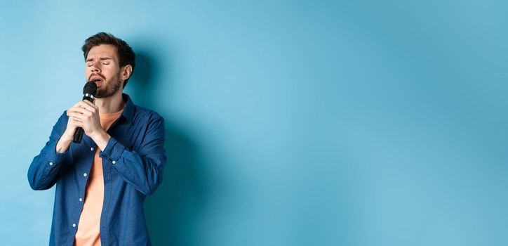 Romantic guy singing in microphone, performing on stage, standing on blue background.