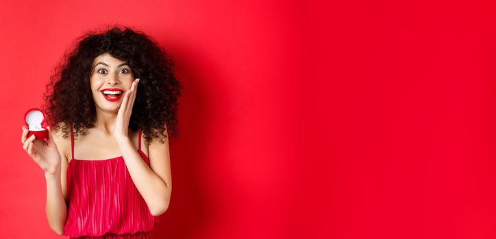 Image of surprised young woman with curly hairstyle, wearing red dress and lipstick, showing engagement ring, going to get married, standing on studio background.