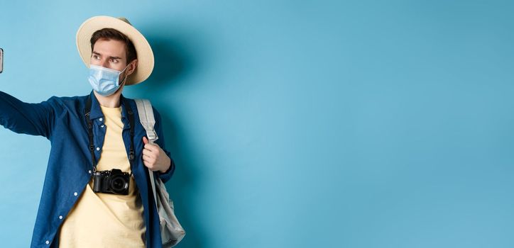 Covid-19, pandemic and travel concept. Tourist on summer vacation taking selfie in medical mask, photographing on smartphone, standing on blue background.