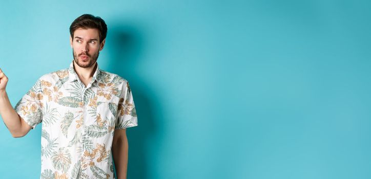 Handsome guy wearing summer shirt on vacation, pointing and looking aside at empty space, making amazed face, standing on blue background.