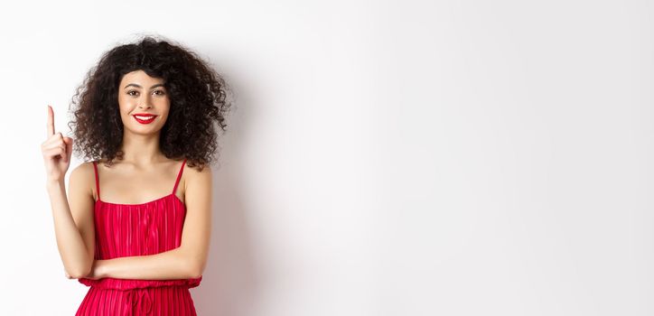 Beautiful smiling lady in red dress showing number one, raising finger and looking pleased, standing over white background.