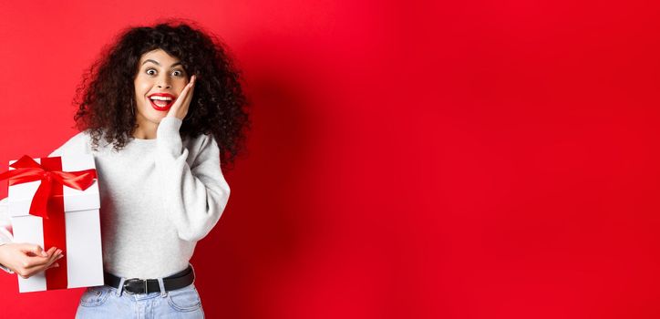 Surprised pretty girl receive Valentines day romantic gift, holding present box and looking with disbelief and happiness at camera, standing on red background.