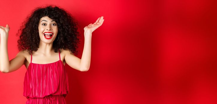 Excited beautiful woman raising hands up and scream with happiness and joy, applause with rejoice, standing on red background.