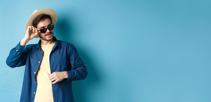 Handsome tourist in sunglasses and straw hat looking aside, smiling pleased, exploring sighsteeing on vacation, blue background.