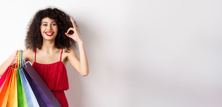 Beautiful woman in elegant dress, holding shopping bags and showing okay sign, recommend store with discounts, white background.