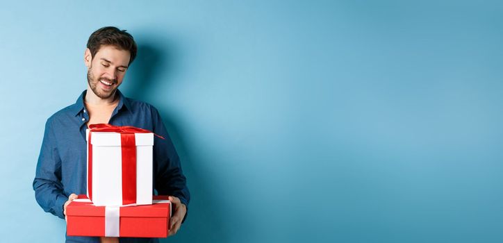 Romantic young man with beard, looking happy at gift boxes on valentines day, giving presents to lover, standing over blue background.